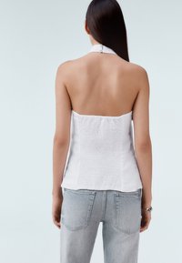 Fehér pántos felső sima elülső résszel és texturált hátsó panellel, világoskék farmernadrággal párosítva, testhezálló kialakítást bemutatva.