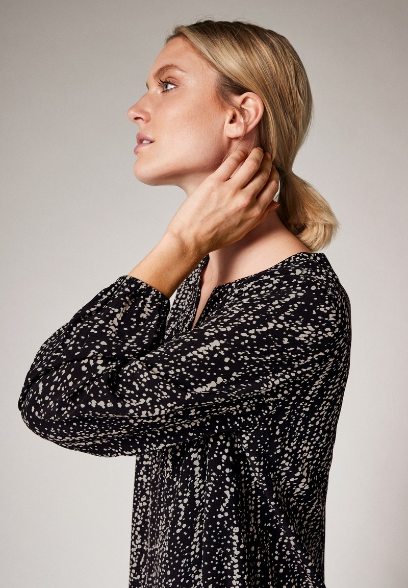 Schwarze Bluse mit einem weißen Spritzmuster, mit langen, gepufften Ärmeln und einem V-Ausschnitt, seitlich gegen einen neutralen Hintergrund gezeigt.