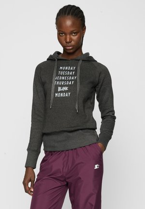 Žena v tmavosivej mikine s textom dní v týždni a fialových športových nohaviciach s bielym logom hviezdy, stojaca pred jednofarebným pozadím.