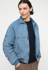 Jeans jakna svijetloplave boje s patentnim zatvaračem sprijeda, rebrastim manžetama i džepom na prsima. Nosena preko tamnoplave puloverice s urezanim natpisom.