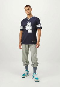 Maglietta da football della Marina con accenti grigi e bianchi, con il numero 4. Abbinata a pantaloni della tuta grigi e scarpe da ginnastica blu e bianche.