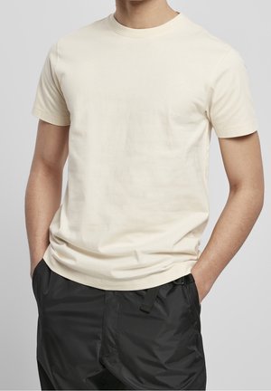 Man draagt een effen off-white T-shirt met korte mouwen met handen in de zakken van losse zwarte broek tegen een effen grijze achtergrond.
