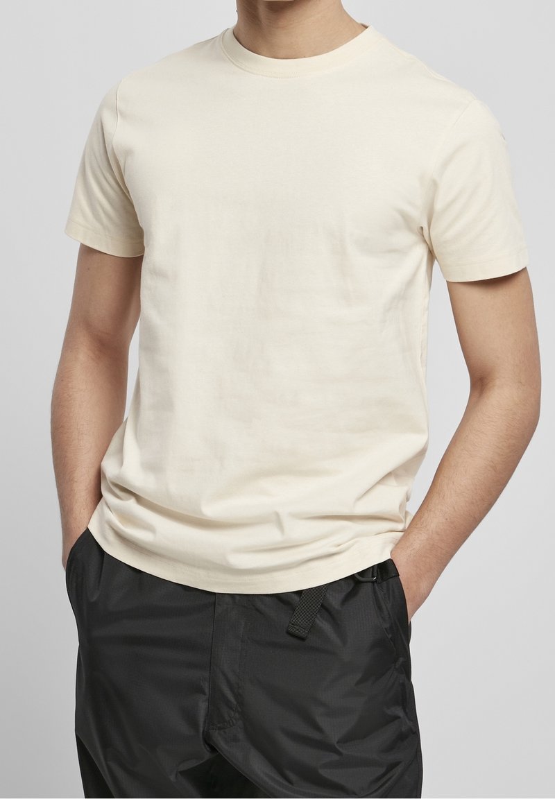 Homme portant un t-shirt uni à manches courtes blanc cassé avec les mains dans les poches d'un pantalon noir ample, sur un fond gris uni.