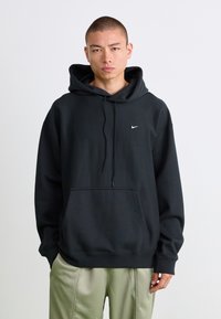 Ung mann med kort hår iført en svart Nike hettegenser og lysegrønne bukser, stående mot en enkel hvit bakgrunn.