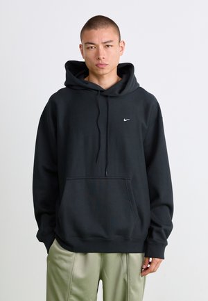 Jonge man met kort haar, gekleed in een zwarte Nike hoodie en lichtgroene broek, staand tegen een simpele witte achtergrond.