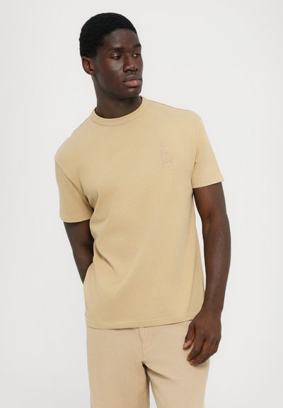 Beige bomulls t-shirt med rund halsringning och korta ärmar. Har en subtil broderad logotyp på bröstet. Slät textur, avslappnad passform.