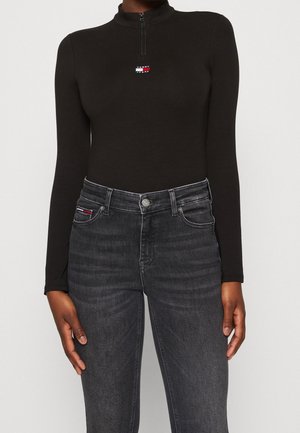 Personne portant un haut noir cintré à manches longues avec une fermeture éclair sur la moitié et le logo Tommy Jeans, associé à un jean noir délavé taille haute.