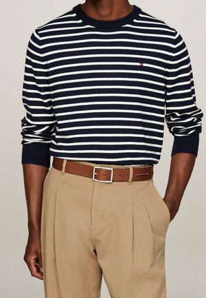 Homme portant une chemise à manches longues rayée marine et blanche rentrée dans un pantalon plissé beige avec une ceinture marron, main dans la poche.