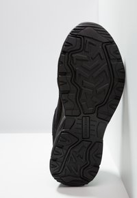 Semelle de chaussure de sport noire avec un motif de semelle robuste pour une meilleure adhérence, en caoutchouc avec une finition texturée et un label de marque visible.