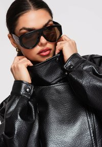 Mujer con grandes gafas de sol negras y una chaqueta de cuero negra con textura, ajustándose el cuello con ambas manos.