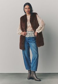Bruine nepbont vest over een doorschijnend, gemusterd wit blouse en blauwe high-waisted jeans, aangevuld met bruine enkellaarsjes.