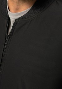 Giacca bomber nera con texture liscia, chiusura con cerniera e un colletto grigio a maglia discreto. Il materiale è leggero e versatile.