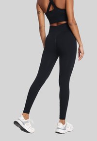 Kobieta stojąca w czarnych legginsach i biustonoszu sportowym w białych sneakersach, widok z tyłu na jednolitym szarym tle.