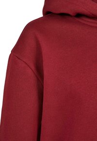 Detailaufnahme eines roten Kapuzenpullis, die die Stoffstruktur, die Schulternaht und die Kapuze zeigt, die hinten liegt.