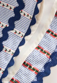 Tissu rayé bleu et blanc avec petits motifs floraux rouges et verts, bordé d'un galon ondulé bleu foncé sur fond de tissu beige clair.