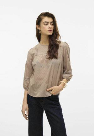 Blouse marron clair avec des manches bouffantes, un tissu texturé et une finition en dentelle le long de l'encolure. Associée à un jean en denim foncé.