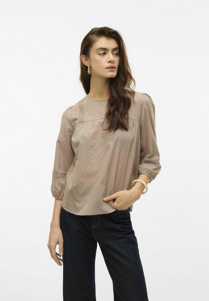 Blusa marrone chiaro con maniche a palloncino, tessuto strutturato e accento in pizzo lungo il collo. Abbinata a jeans scuri in denim.