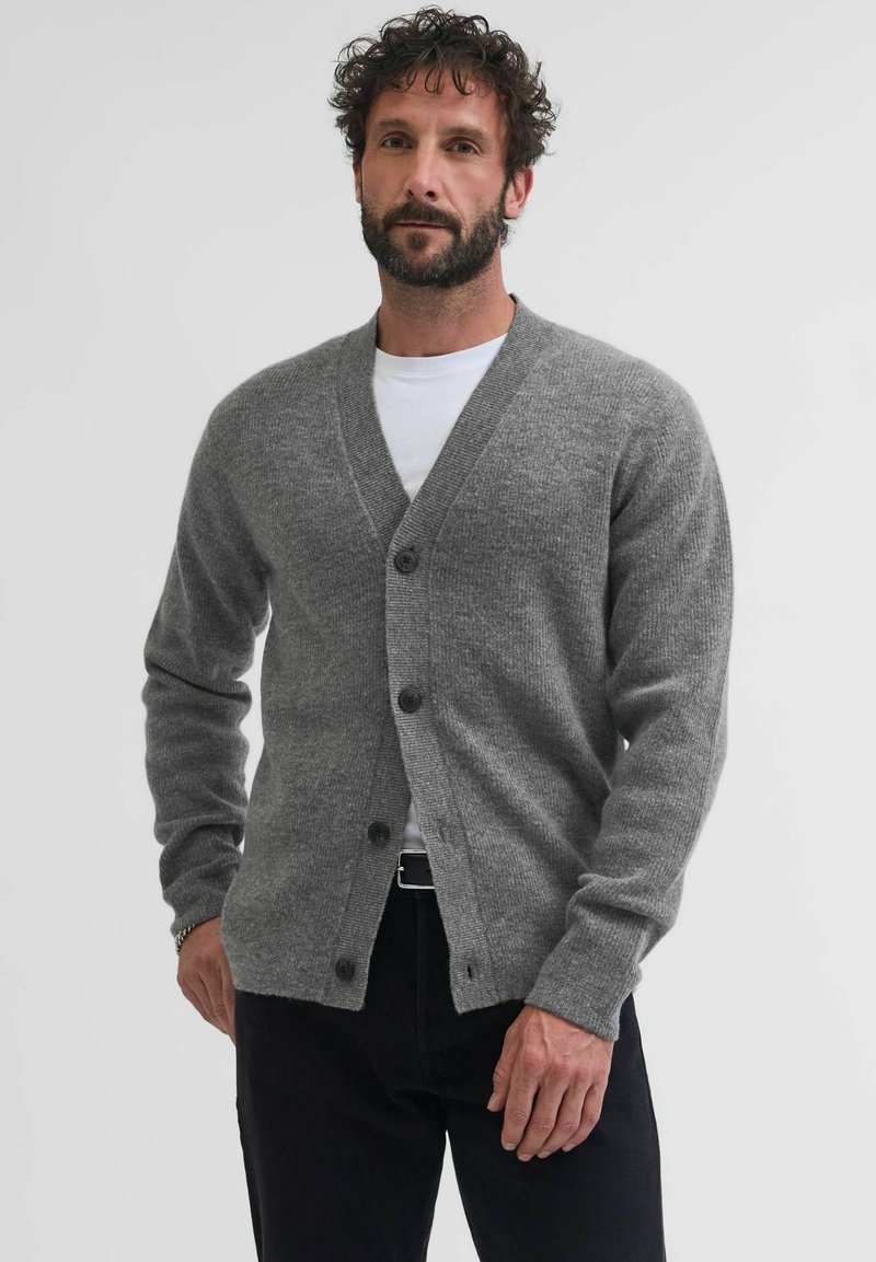 Cardigan de lana gris con escote en V, cinco botones y mangas largas. Presenta una textura suave y un ajuste relajado. Se lleva sobre una camisa blanca.