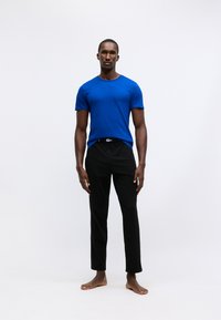 T-shirt da uomo blu in cotone con maniche corte abbinato a pantaloni neri. I pantaloni presentano una cintura nera. Il modello è in piedi a piedi nudi su uno sfondo neutro.