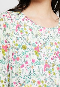 Blouse fleuri avec un fond blanc et une variété de fleurs colorées en rose, jaune et vert. Tissu doux, col rond.