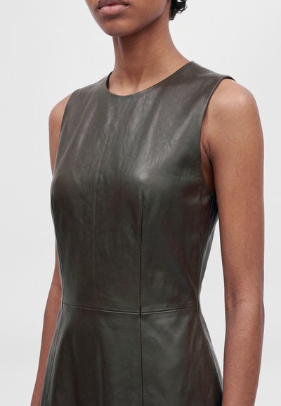 Modèle portant une robe en cuir marron foncé, ajustée et sans manches, avec une encolure ronde et une couture de taille apparente.