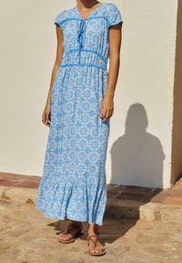 Niebieska maxi sukienka w wzory z krótkimi rękawami i marszczonym pasem; ma detal w postaci wiązania przy dekolcie oraz falbaniasty dół. Noszona z brązowymi sandałami.