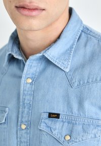 Jasnoniebieska koszula z denimu z wyprofilowanym kołnierzykiem, dwiema kieszeniami na piersi, srebrnymi zatrzaskami oraz czarną metką "Lee" na górnej kieszeni.