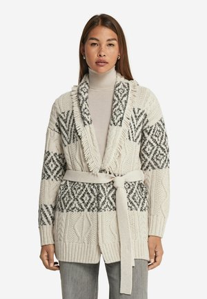 Strikjakke i cremefarvet med sorte geometriske mønstre, med sjalkrave, bælte i taljen og ribbede manchetter. Tekstur i kabelstrik.
