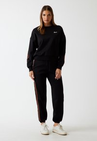 Completo felpa nera con pantaloni, caratterizzato da strisce laterali marroni. Realizzato in materiale morbido, presenta un collo rotondo e polsini affusolati. Logo bianco sulla felpa.
