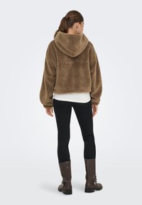 Bruine fleece cropped hoodie met een capuchon, textiel met structuur, oversized pasvorm en verlaagde schouders, gecombineerd met zwarte spijkerbroek en bruine laarzen.
