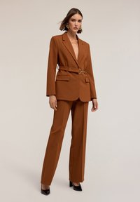 Traje marrón a medida con cintura ceñida, con un escote profundo en forma de V y bolsillos de solapa. Combinado con zapatos de tacón altos negros.