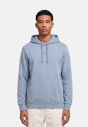 Giovane uomo che indossa una felpa con cappuccio Puma azzurro chiaro e pantaloni beige, in piedi davanti a uno sfondo bianco uniforme.