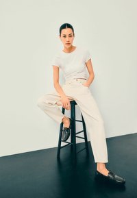 Lichtbeige, hoog-waardige wijde broek in combinatie met een getailleerd wit T-shirt en zwarte bootschoenen, zittend op een zwarte kruk.