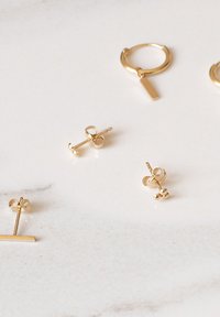 Gouden oorbellen op een lichte marmeren ondergrond, met ontwerpen van hoepels, staven en stud oorbellen in verschillende texturen en vormen.