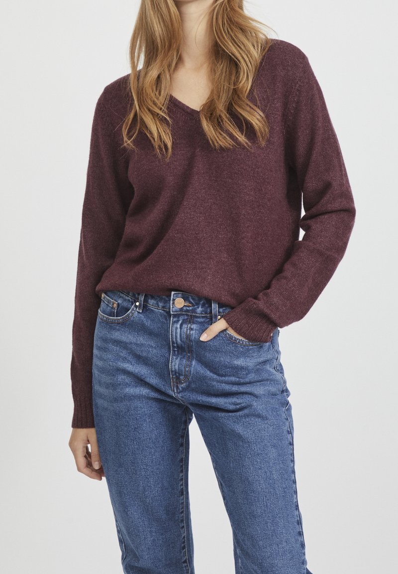 Femme portant un pull col en V bordeaux rentré dans un jean bleu taille haute, une main dans la poche, sur un fond uni.