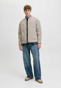 Veste beige matelassée avec de grandes poches avant, col arrondi et fermetures à pression, portée sur une chemise à carreaux et associée à un jean ample.