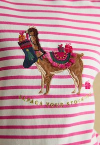 Biela tkanina s ružovými pruhmi, na ktorej je vyšitá hnedá lama s ponožkou a darčekmi, doplnená dekoratívnymi akcentmi a textom pod ňou.