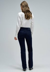 Mujer con cabello largo y ondulado, vistiendo una blusa blanca y jeans azul oscuro, de pie descalza de espaldas frente a un fondo liso.