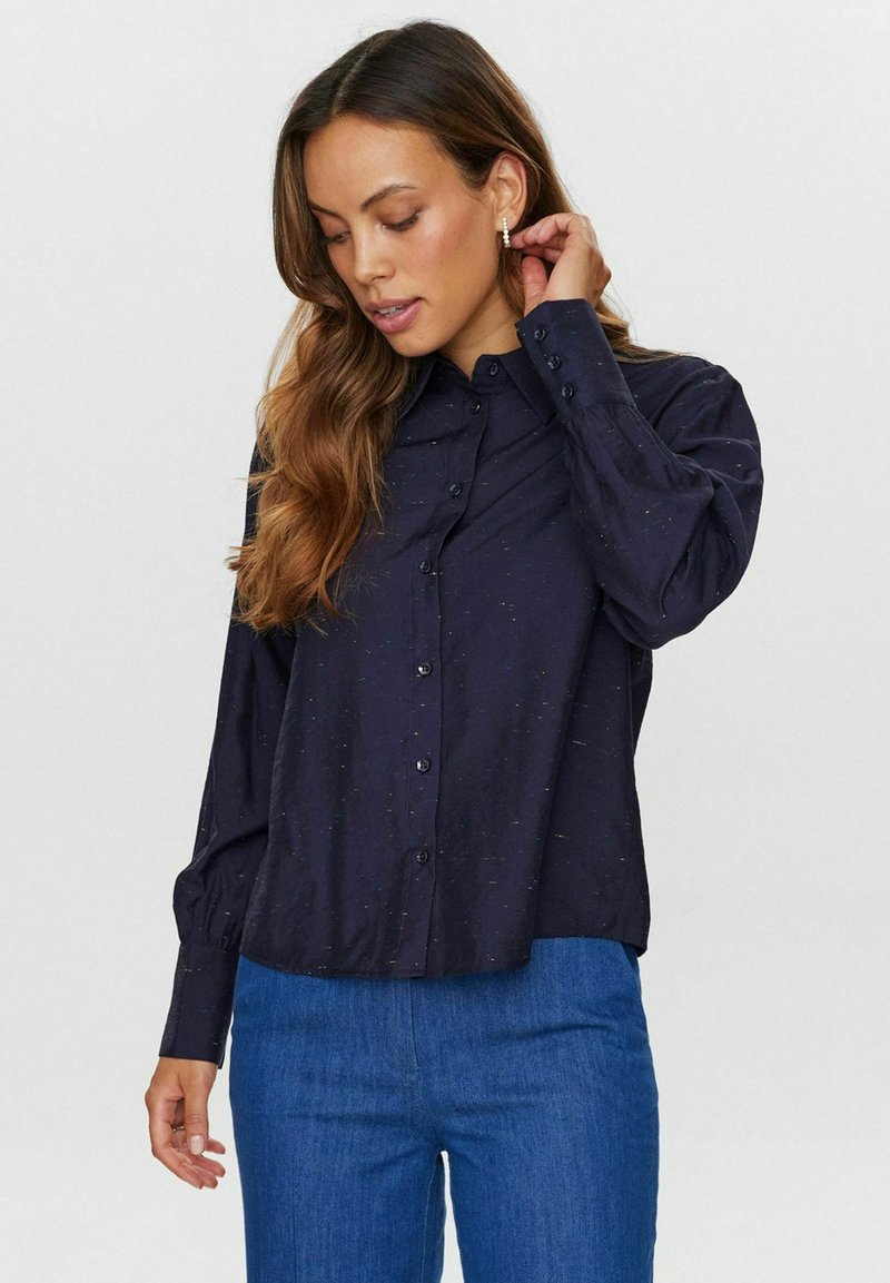 Navy button-up skjorte med krave, lange ærmer og subtilt tekstureret mønster, parret med blå bukser, sat mod en neutral baggrund.