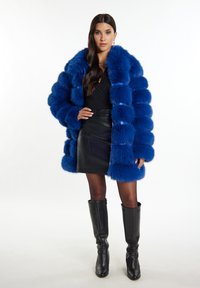 Helle blaue Kunstpelzjacke mit großen, weichen, strukturierten Streifen. Getragen über einem schwarzen Oberteil und einem Lederrock, kombiniert mit hohen schwarzen Stiefeln.