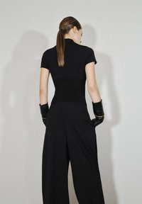 Crni uski top s kratkim rukavima, uparen s širokim crnim hlačama. Model nosi crne rukavice, kosa je vezana, a pozadina je minimalna.