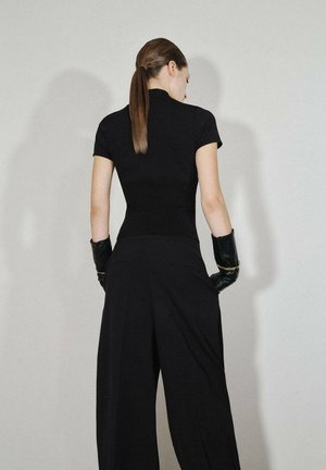 Top negro ajustado de manga corta, combinado con pantalones anchos negros. La modelo lleva guantes negros, el cabello recogido, con un fondo minimalista.