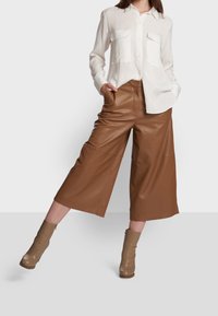 Biała koszula z długim rękawem z dwoma kieszeniami na piersi, połączona z brązowymi skórzanymi spodniami culottes i taupe botkami, umieszczona na neutralnym tle.