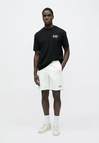 Juoda medvilninė marškinėliai su logotipu, balti šortai, balti sportbačiai ir baltos kulkšnies kojinės. Minimalistinis dizainas, laisvas siluetas, tinkamas kasdieniam dėvėjimui.