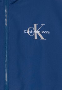 Tamsiai mėlyna striukė su sidabriniu užtrauktuku ir antspaudu su užrašu "Calvin Klein Jeans" bei "CK" priekyje. Glotnus, lengvas audinys.