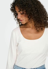 Hvid bluse med lange ærmer og bred rund halsudskæring, glat bomuldsstof, tætsiddende design, og båret med lyseblå denimjeans.