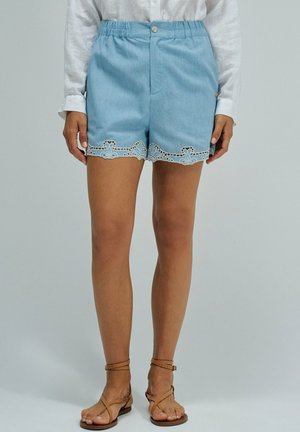 Femme portant un short bleu clair avec un ourlet en dentelle festonnée, un chemisier blanc à manches longues et des sandales plates marron clair à bride à la cheville, debout devant un fond uni.