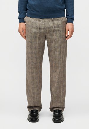 Άνδρας που φοράει καφέ καρό παντελόνι, μαύρα δερμάτινα παπούτσια και μπλε μακρυμάνικη μπλούζα, στέκεται μπροστά σε απλό φόντο.