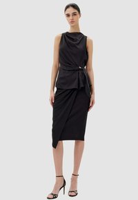 Vestito nero senza maniche con scollo drappeggiato e cintura in vita, con gonna a matita aderente asimmetrica e dettagli brillanti.