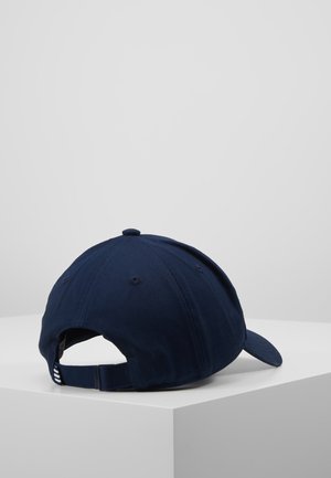 Gorra de béisbol azul marino con correa ajustable, apoyada sobre una superficie blanca, mostrada desde la parte trasera y un ángulo lateral.
